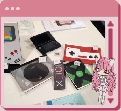 Cadre rose avec Onyx en bas à droite. Papeterie sur le thème rétrogaming : carnets représentants la PS1, la Xbox et la Famicon